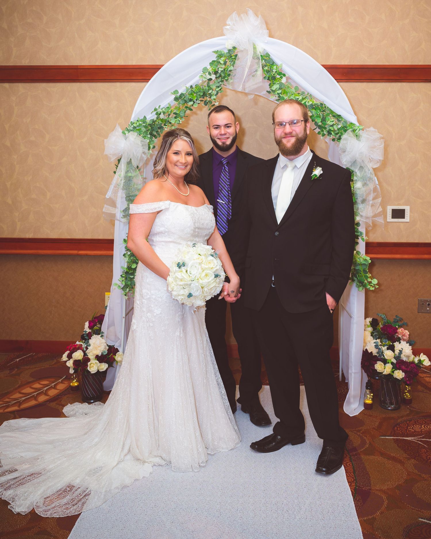 Wedding couple poses with officiant under decorated white arch with greenery and floral accents.