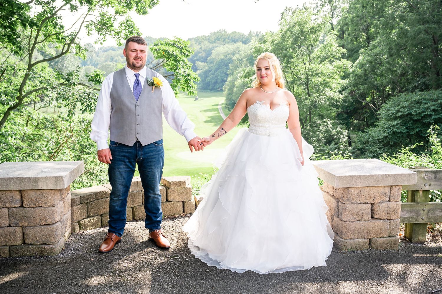 Wedding couple holding hands on stone overlook surrounded by lush greenery.