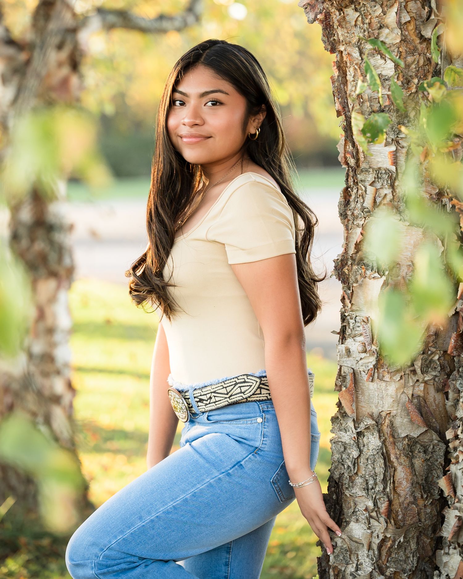 A person in a cream top and jeans poses casually among birch trees in a sunlit outdoor setting.