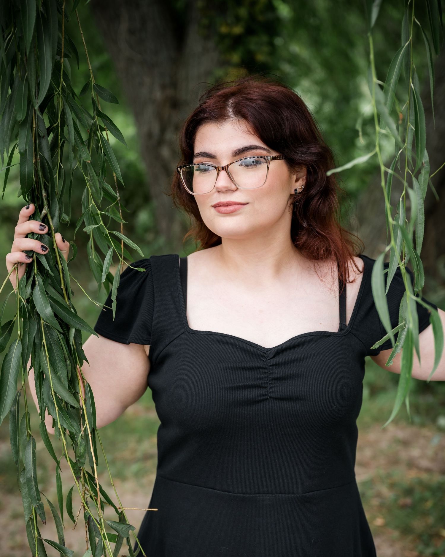 Portrait between willow branches wearing glasses and a black dress.