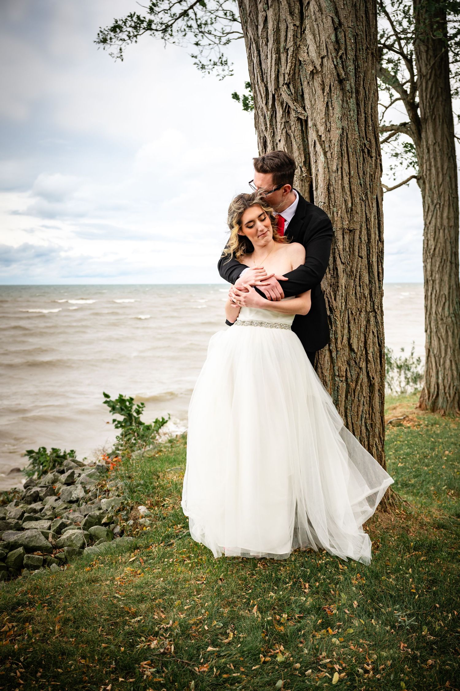 Romantic lakeside wedding portrait with dramatic cloudy sky and large tree backdrop.