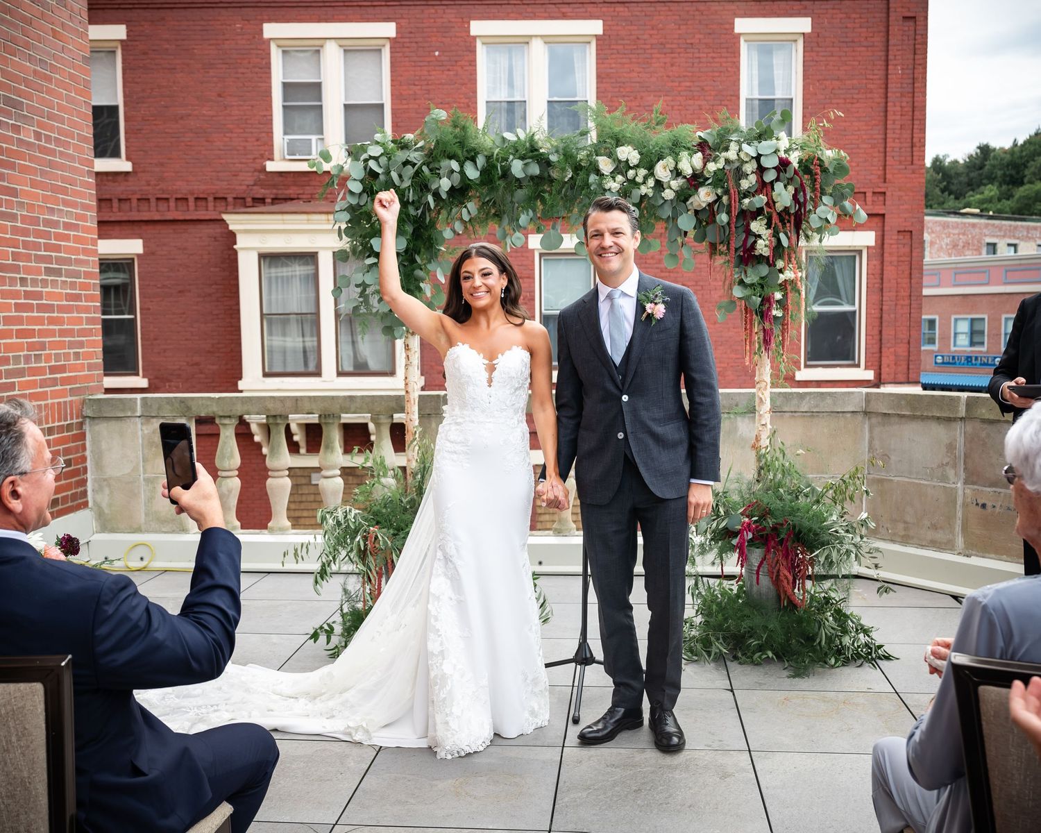 Wedding ceremony under floral arch against brick building backdrop.