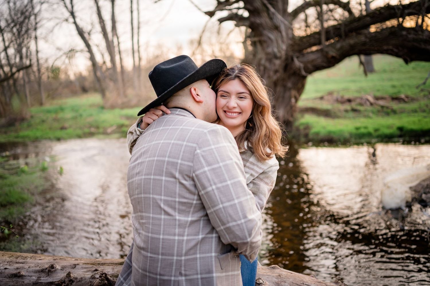 A couple embraces by a tranquil stream on an early spring evening, one wearing a plaid jacket and fedora hat.