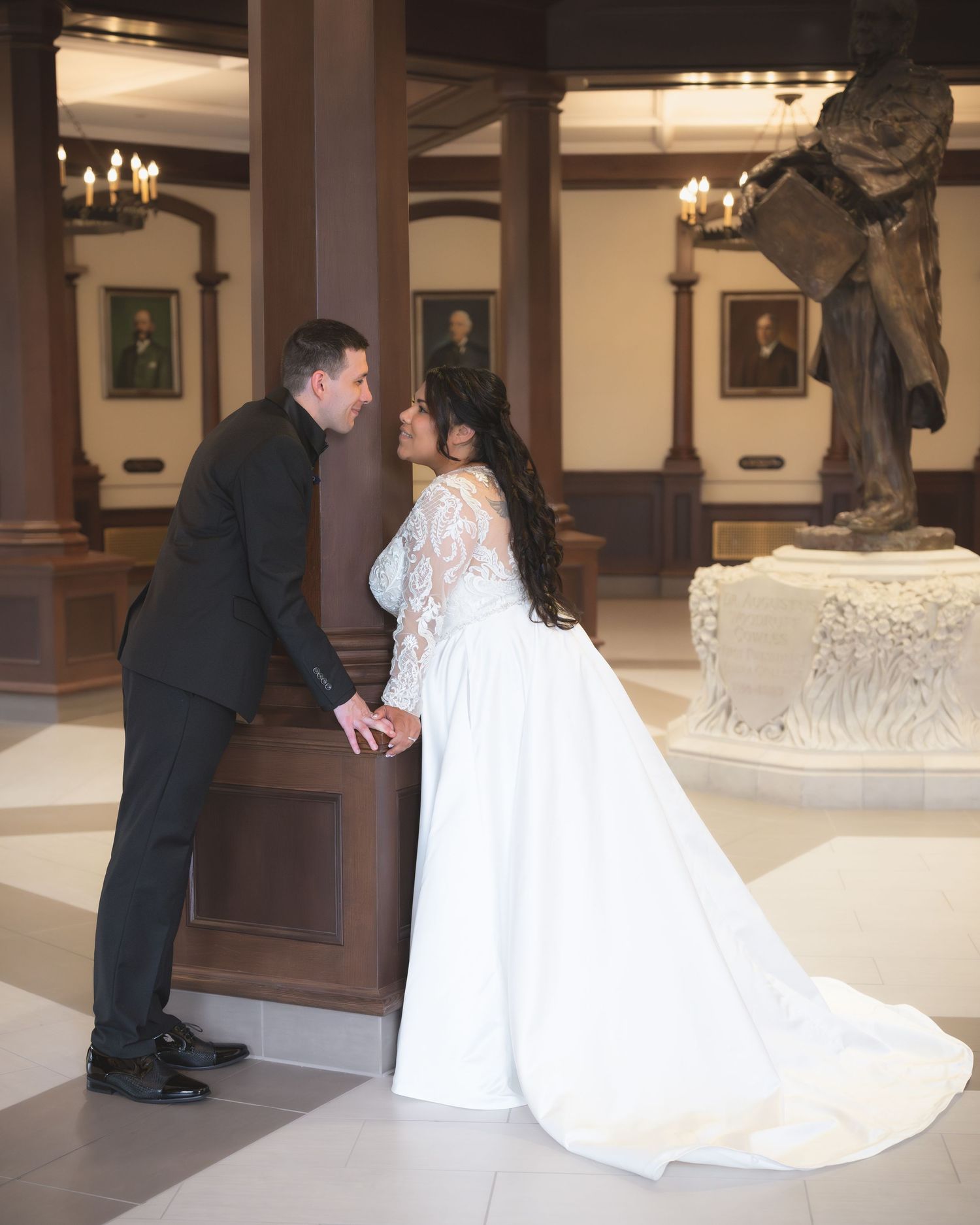 A couple shares an intimate moment in an elegant hall with ornate decor and a grand sculpture nearby.