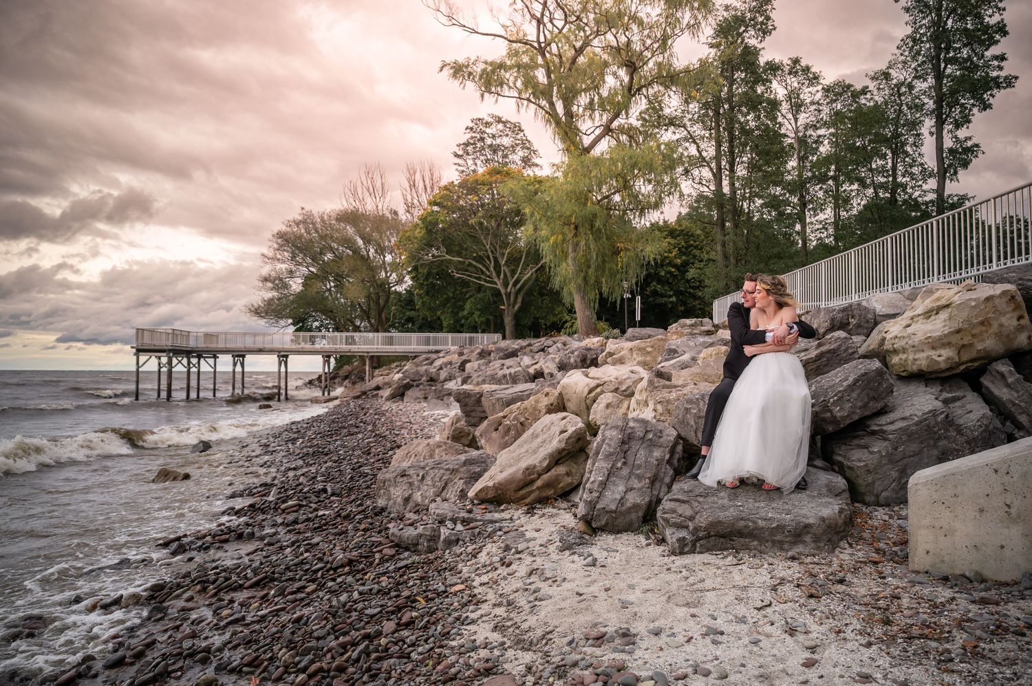 Waterfront wedding portrait with wooden pier and rocky shoreline at sunset.