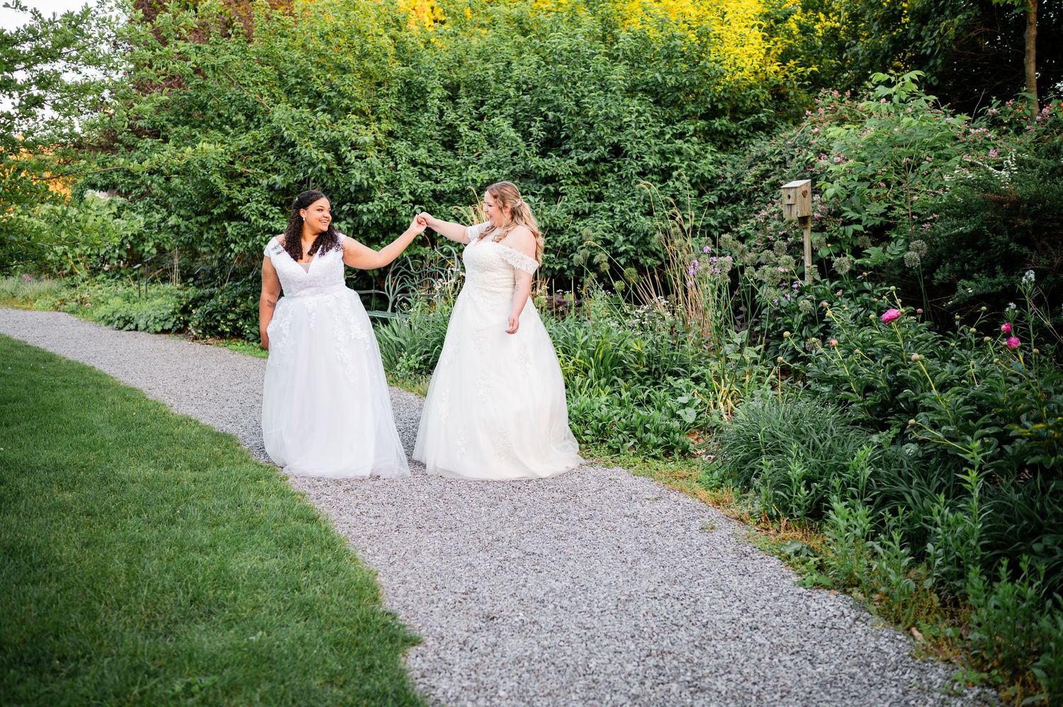 Two brides in white wedding dresses walk together on a garden path surrounded by lush greenery.