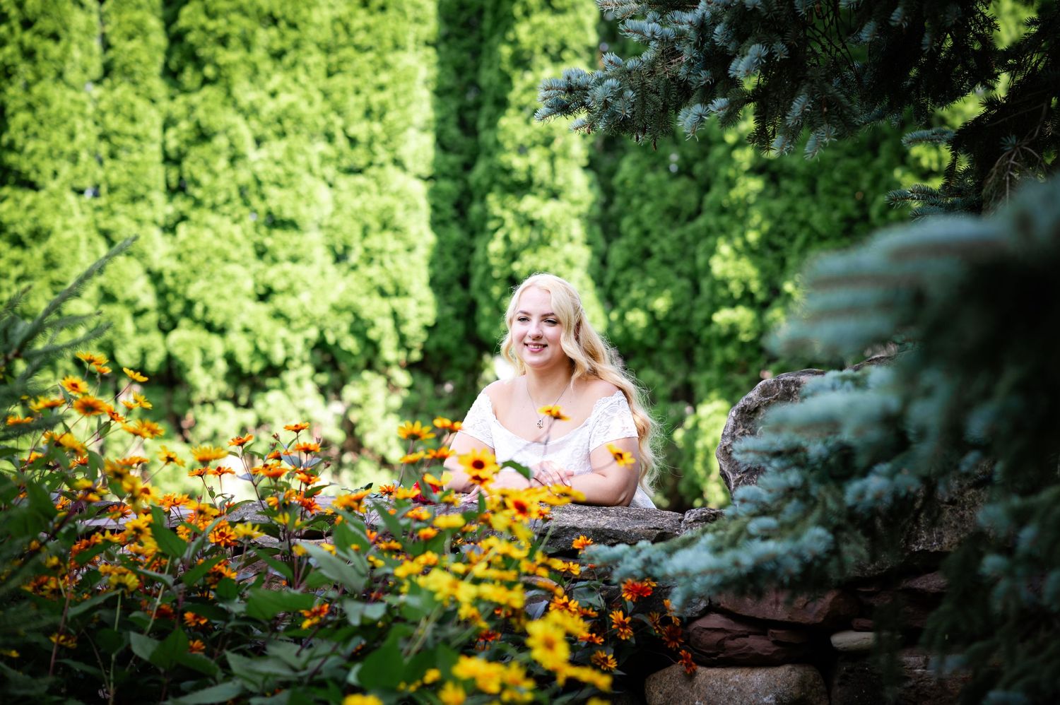 Person in light dress sits by garden wall surrounded by vibrant summer blooms.