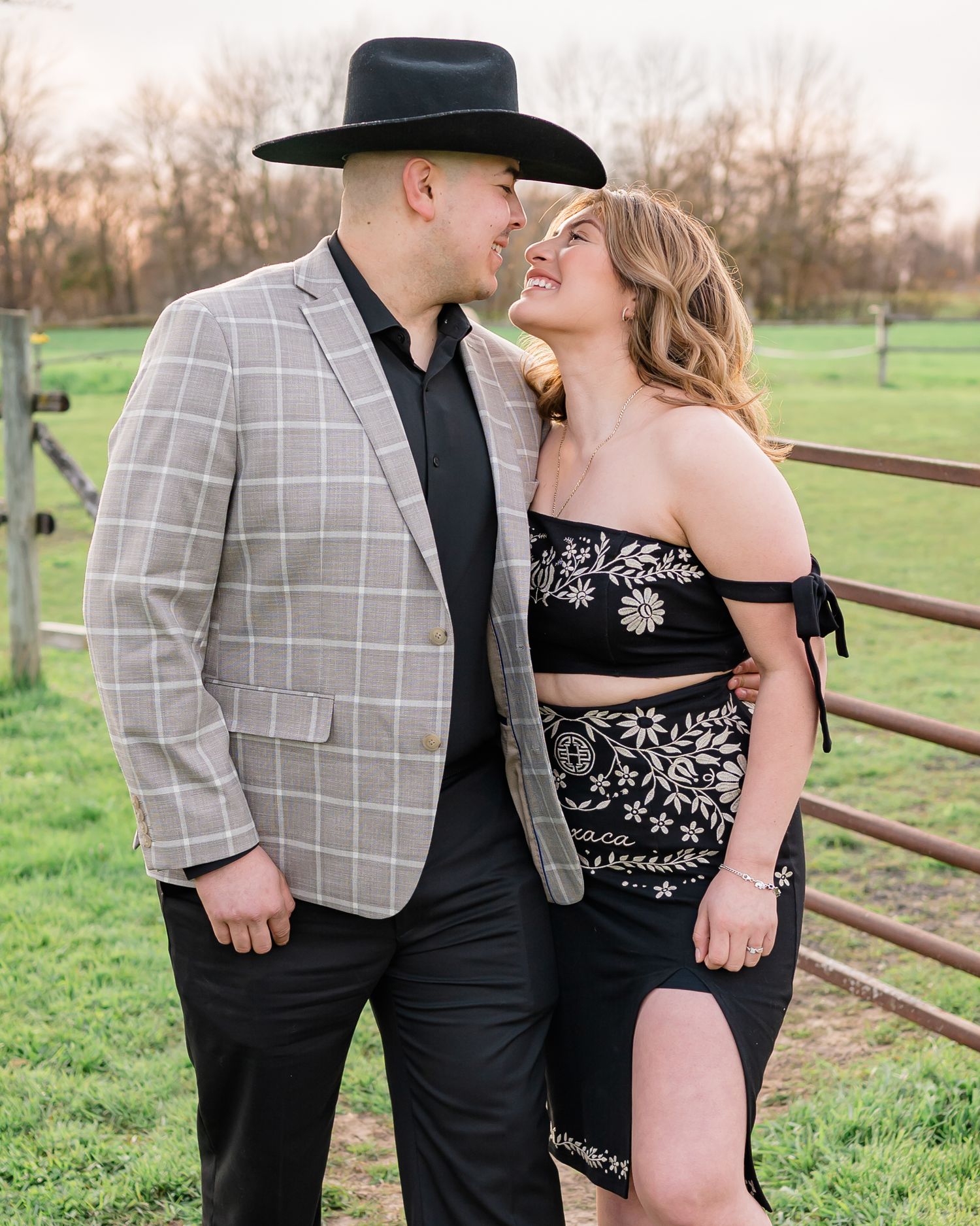 Couple shares intimate moment while posing in western-inspired outfits near wooden fence.