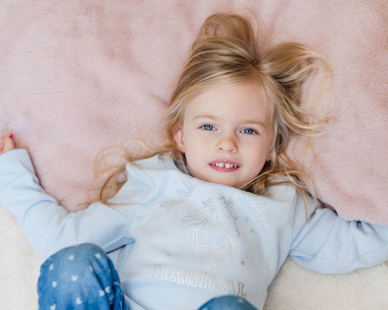 Smiling child lying on a pink blanket wearing a light blue sweater.