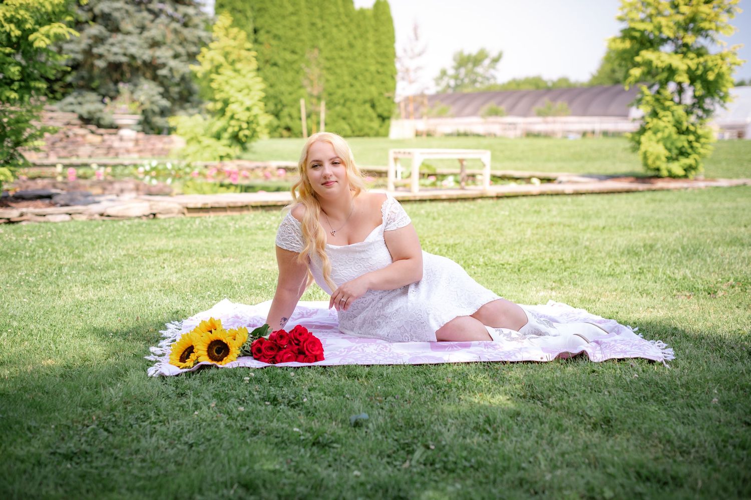 Person in white dress relaxing on blanket with sunflowers and roses in garden.