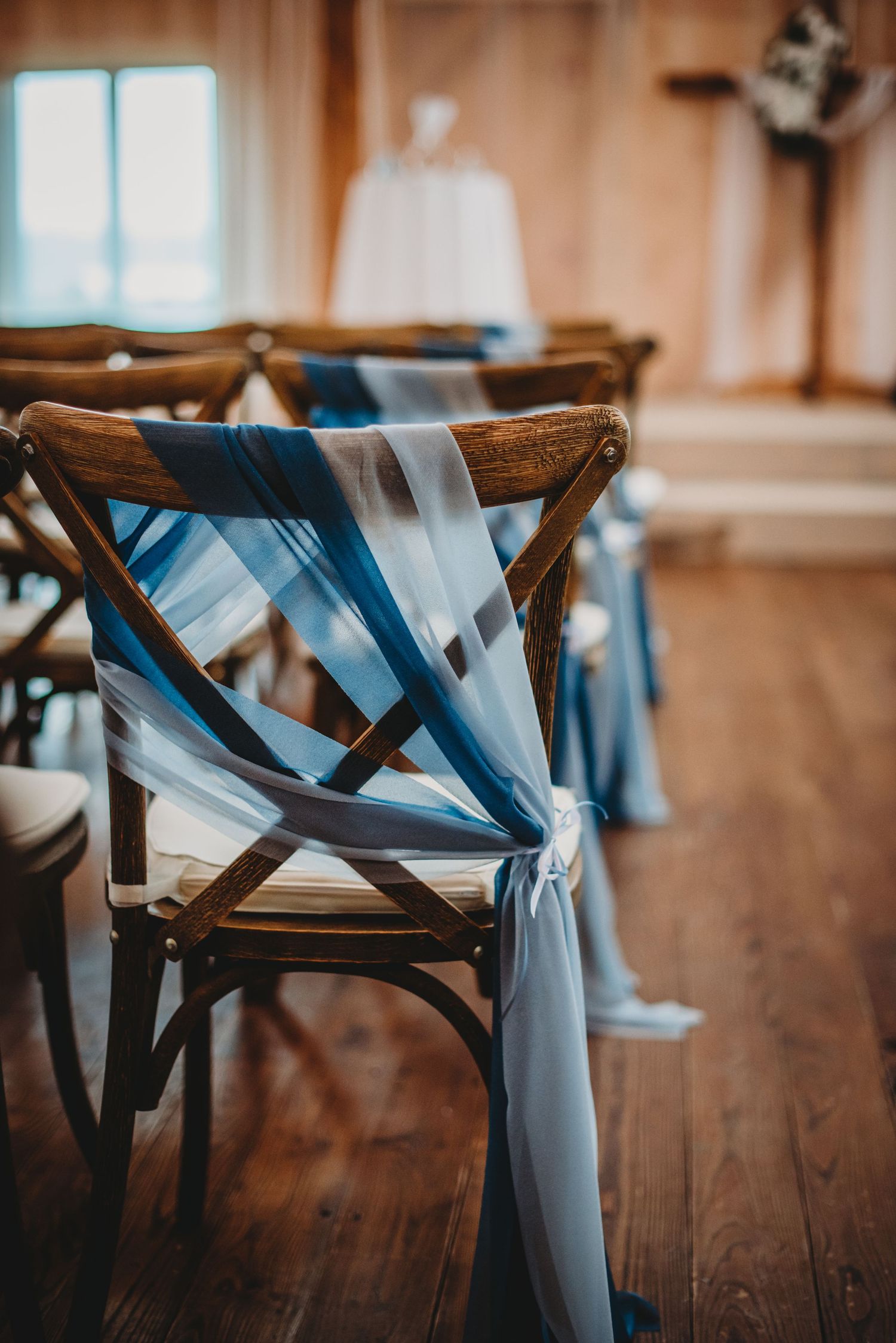 Wooden crossback chairs decorated with flowing blue fabric ribbons in wedding ceremony setup.