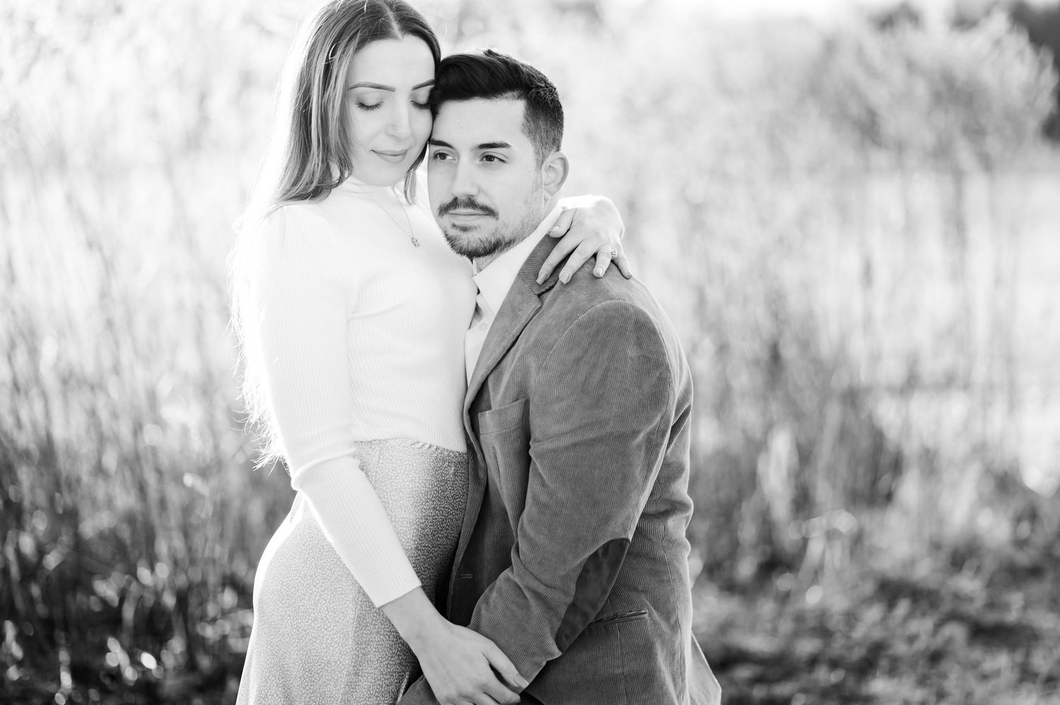 Sweet natural light engagement photo shows couple embracing tenderly in rustic outdoor setting with grass and sunlight.