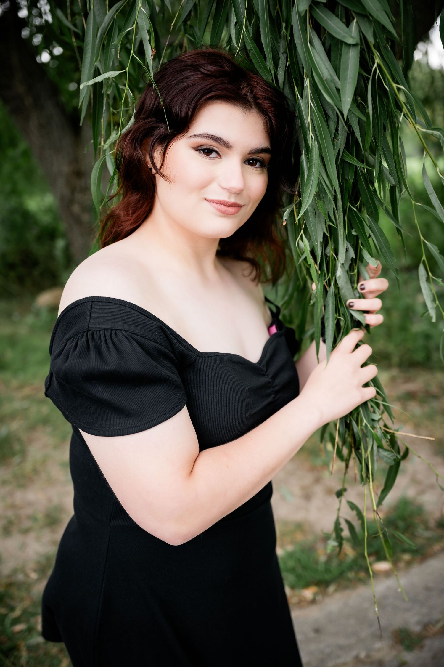 Portrait of woman in black off-shoulder dress posed against hanging willow branches.