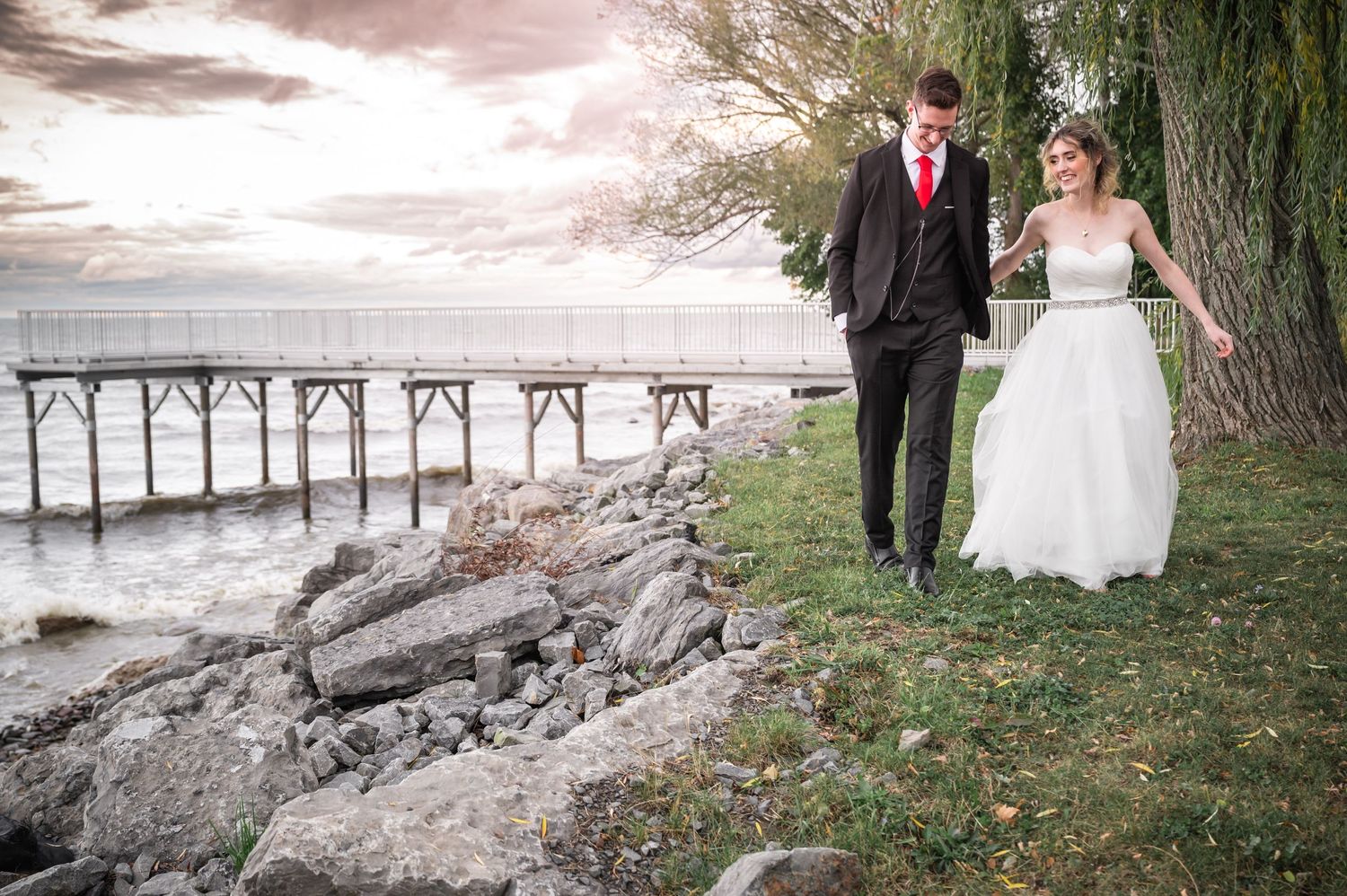 Bride and groom walk along rocky shoreline with vintage pier and dramatic sky backdrop.