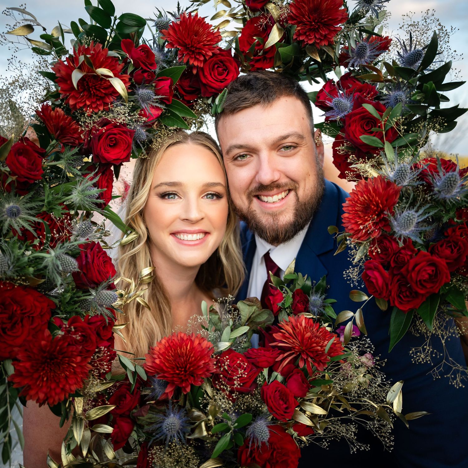 Wedding portrait with deep red floral arrangement of roses and mums framing the couple against a dark background.