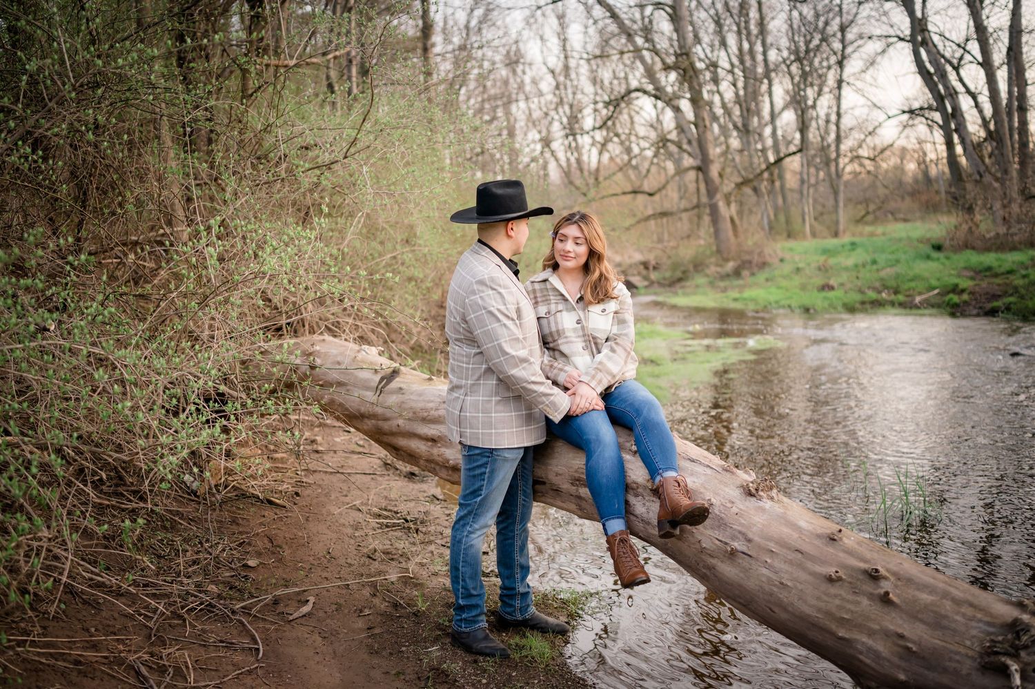 A couple poses together on a log over a peaceful stream in a wooded setting.