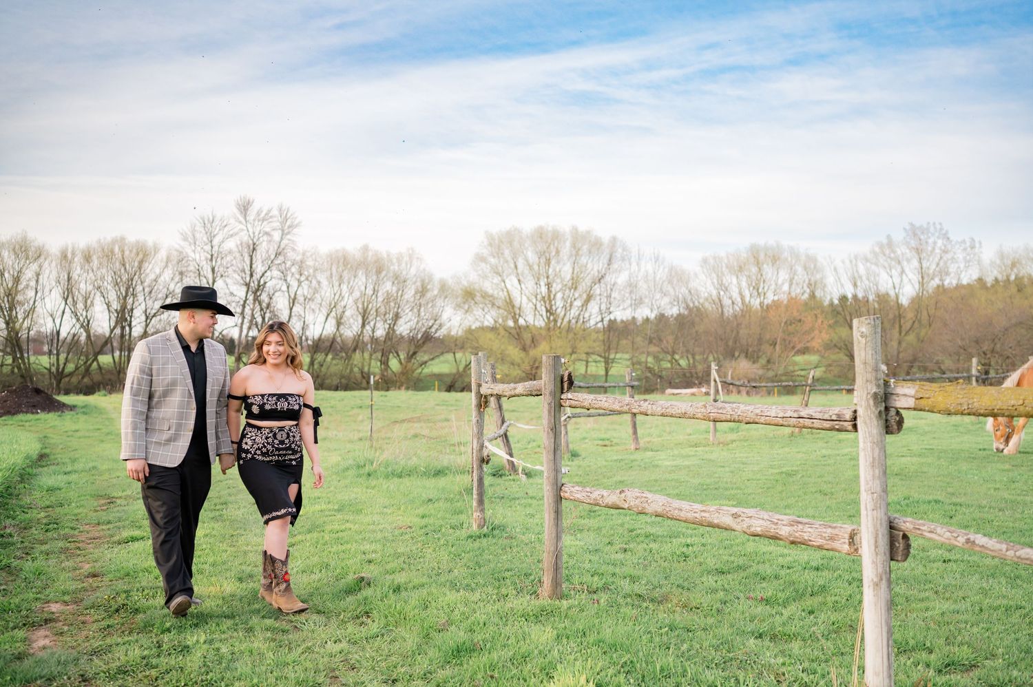 Couple walking along wooden fence line in pastoral setting with cloudy sky above.
