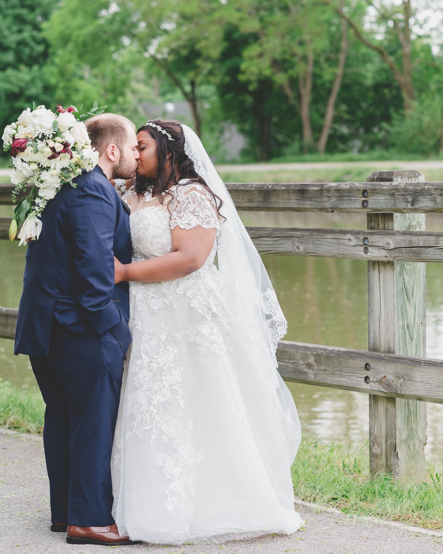 A romantic wedding couple sharing a kiss by a wooden dock over a pond with a beautiful white floral bouquet.