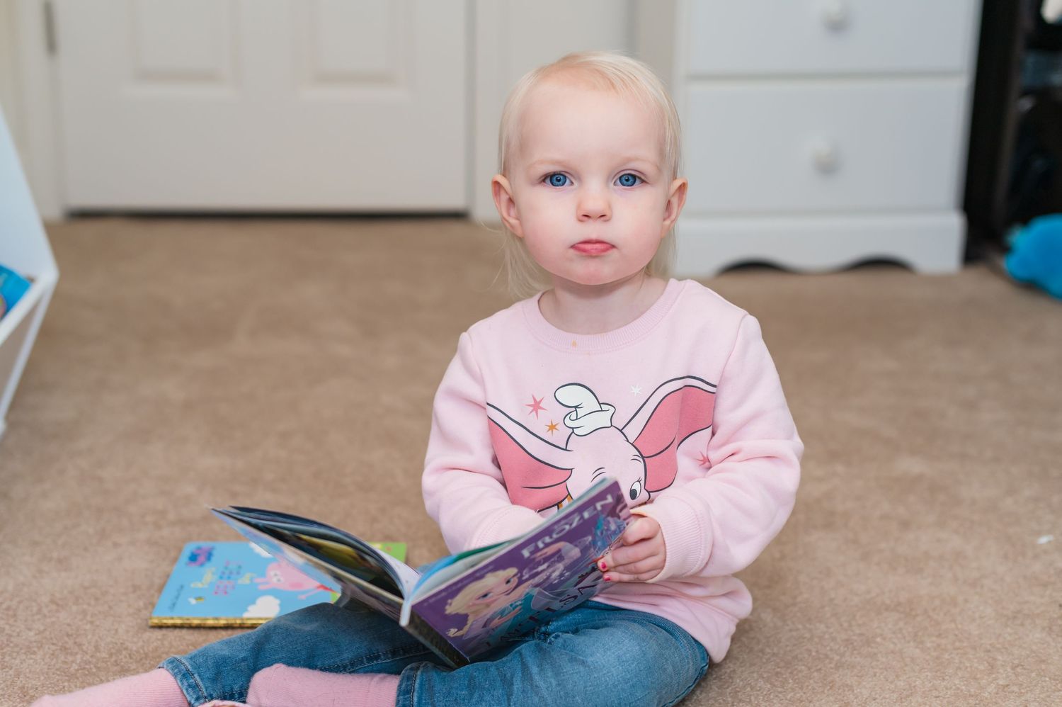 Young child sitting on floor reading a book while wearing a pink sweater.