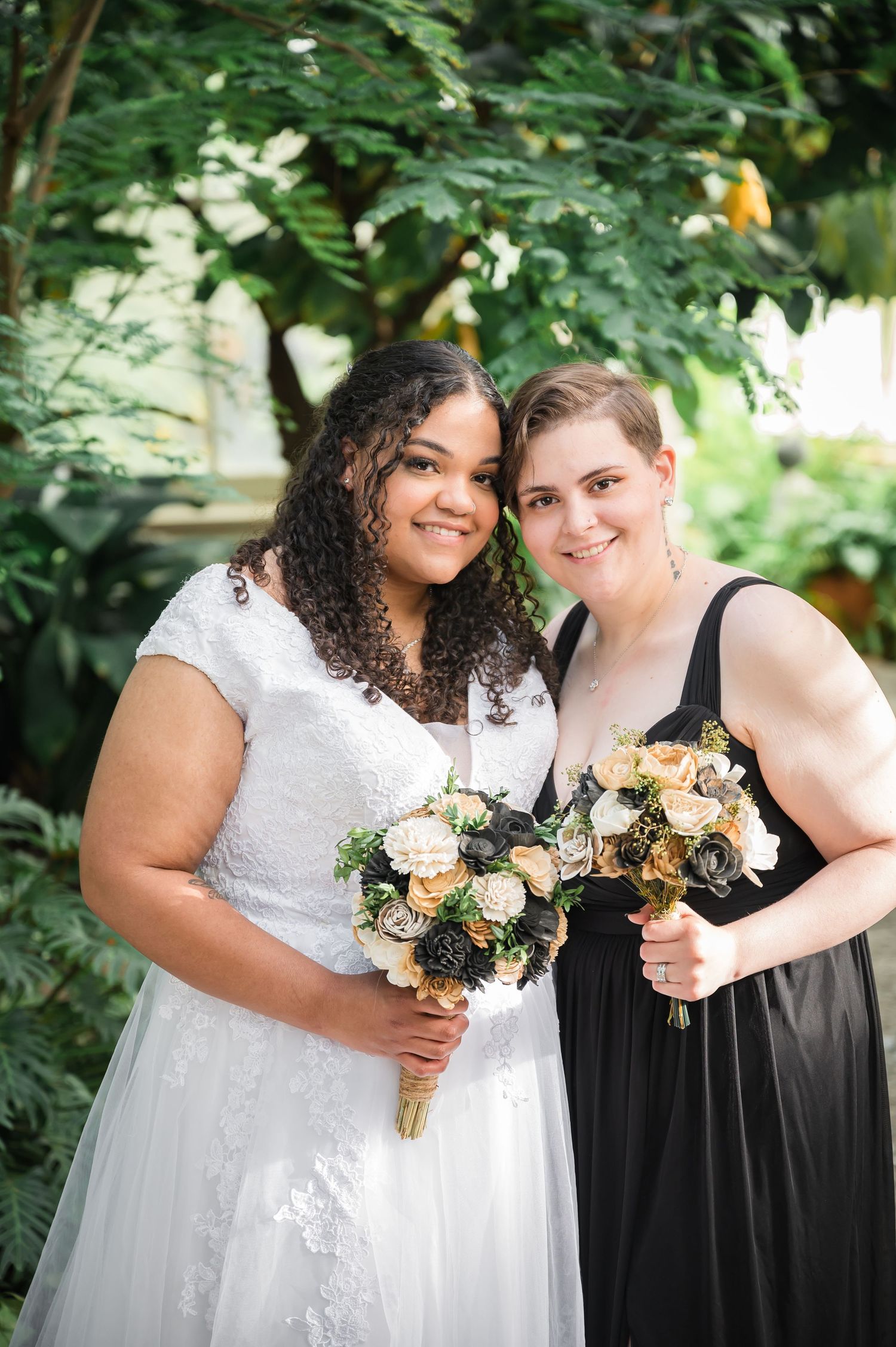 A person in white dress and another in black hold peach and cream floral bouquets in a garden setting.