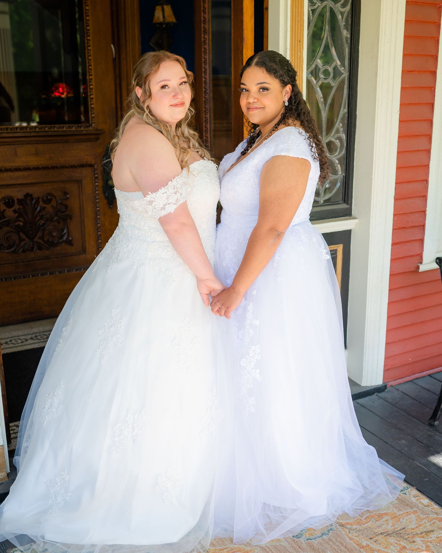 Two people in white wedding dresses pose together on a porch with ornate red door in background.
