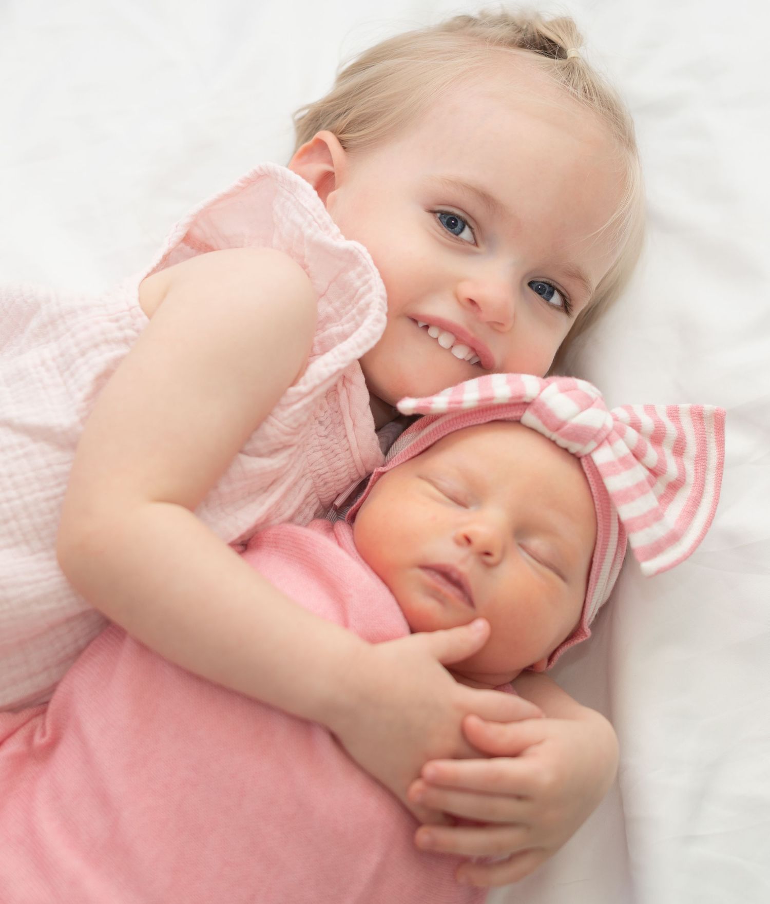 Two children in pink outfits cuddle together on white bedding, with one wearing a striped headband.