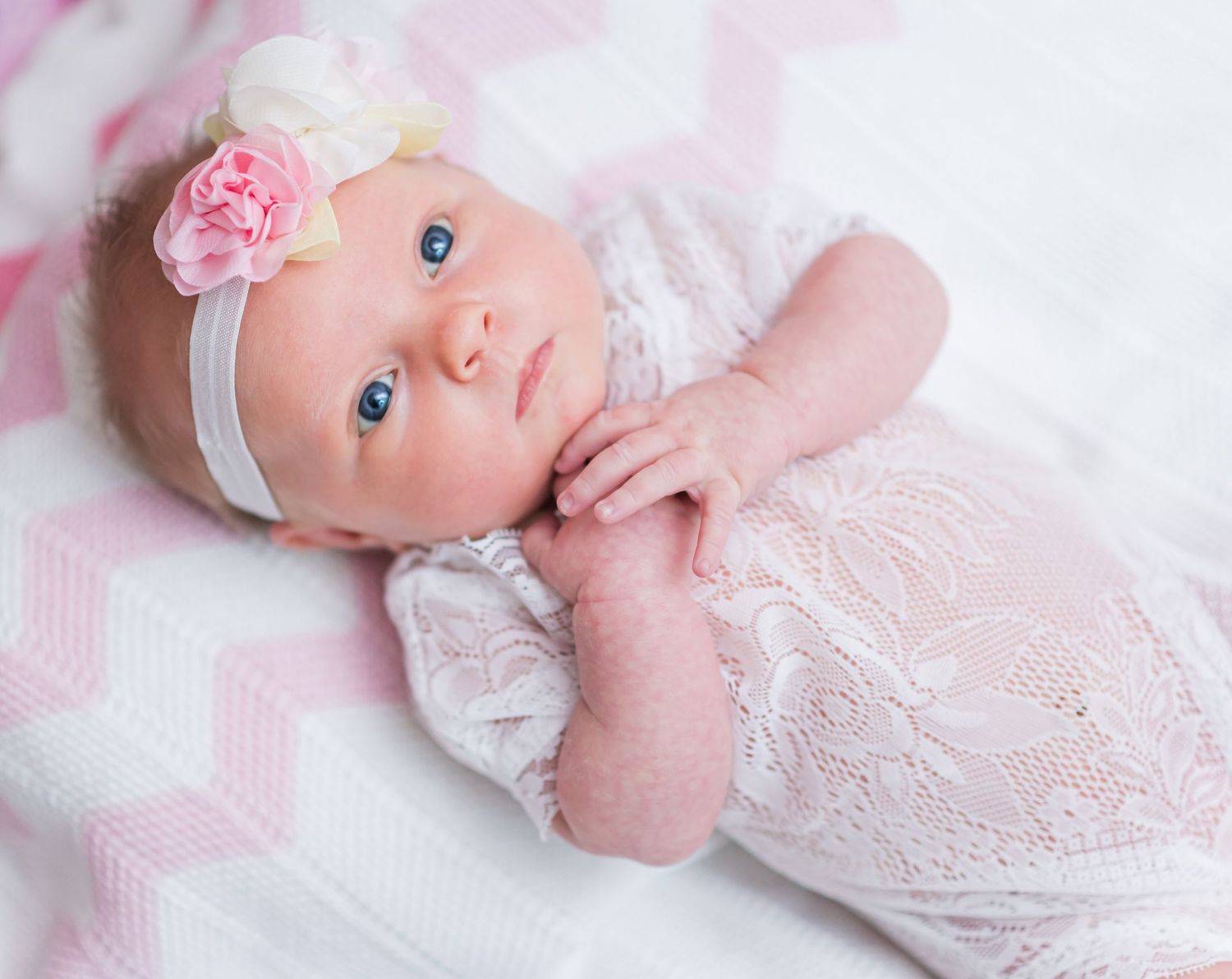 A baby wearing a white lace dress and pink flower headband lies on soft pink fabric.