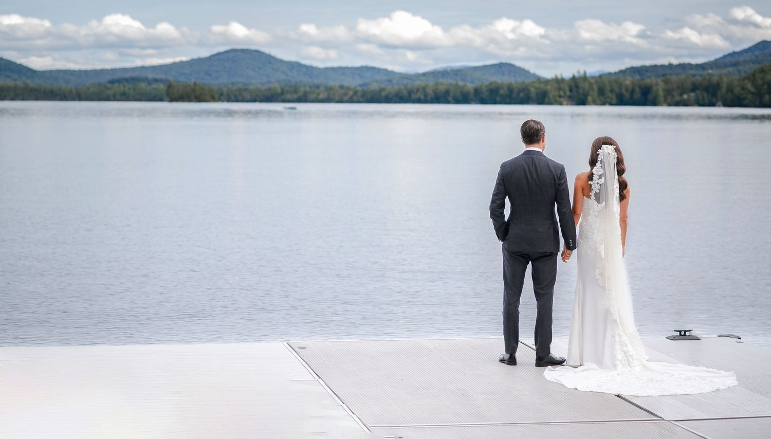 A scenic wedding photoshoot by a serene lake with mountains and forest in the background on a sunny summer day.