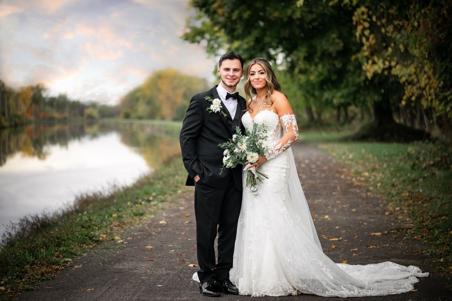 A couple in wedding attire poses on a path by a tranquil river during autumn.