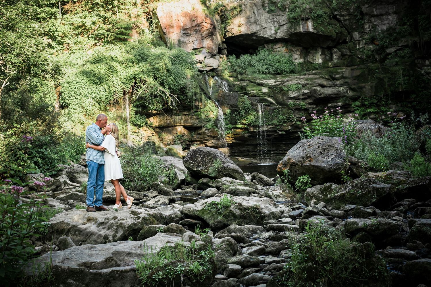 A couple stands together near a scenic waterfall surrounded by rocky terrain and lush greenery.