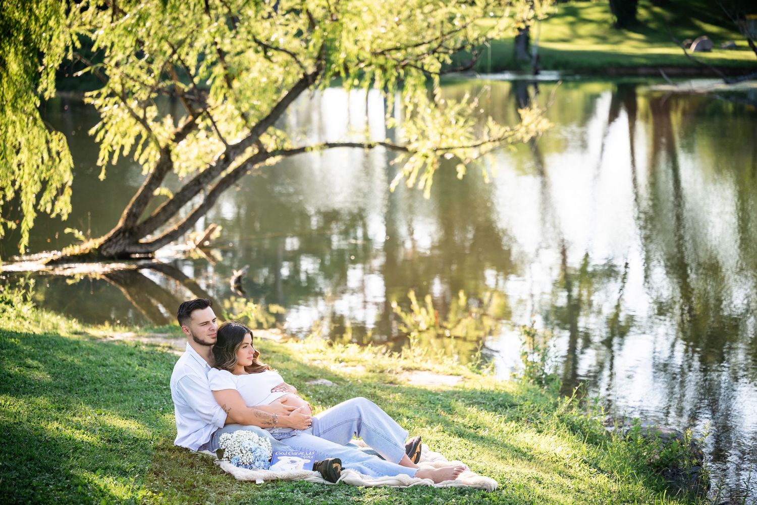 Couple relaxing by a serene pond in a park, woman resting on man's lap amid lush greenery.