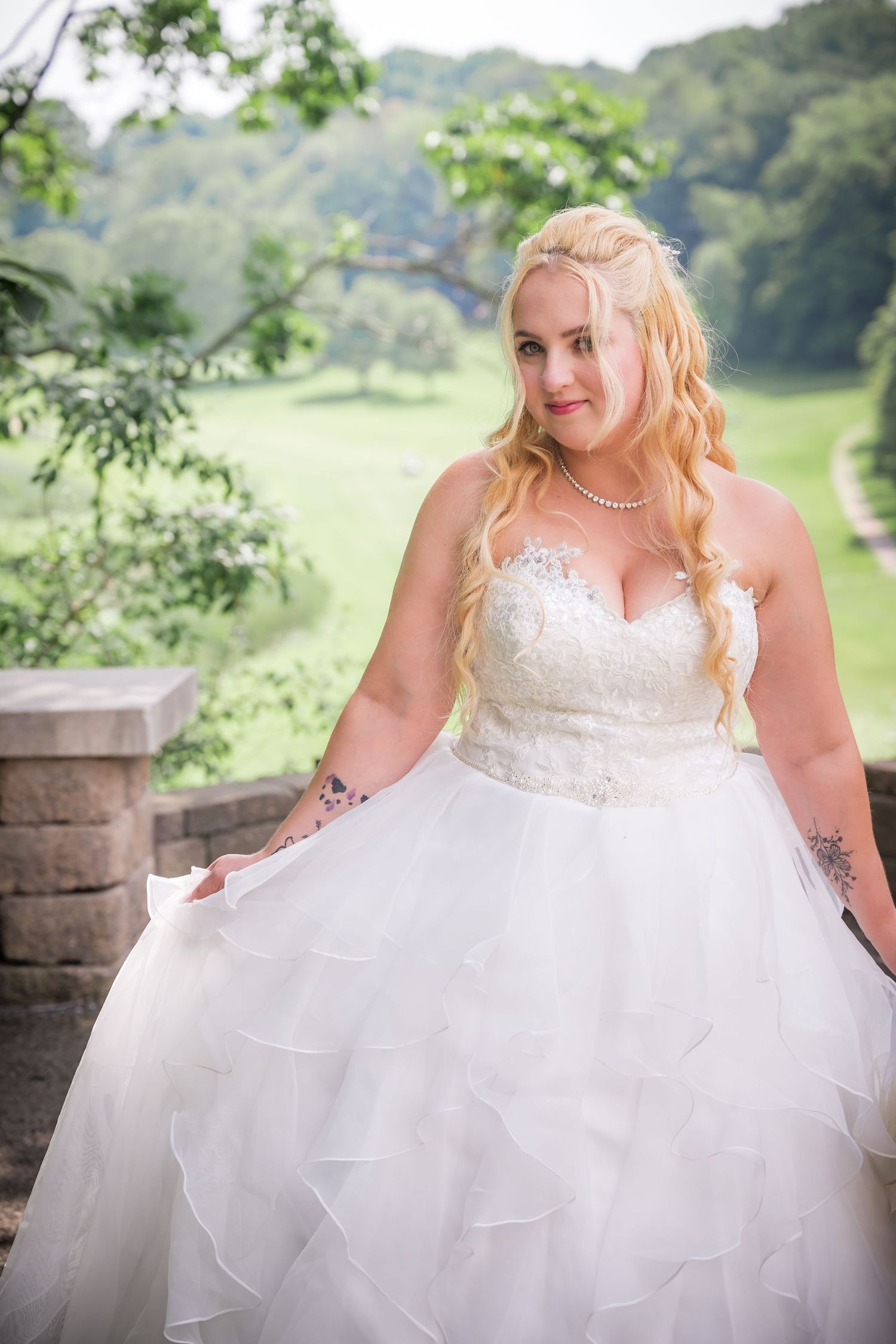 Bride in white strapless gown poses outdoors with scenic overlook in background.