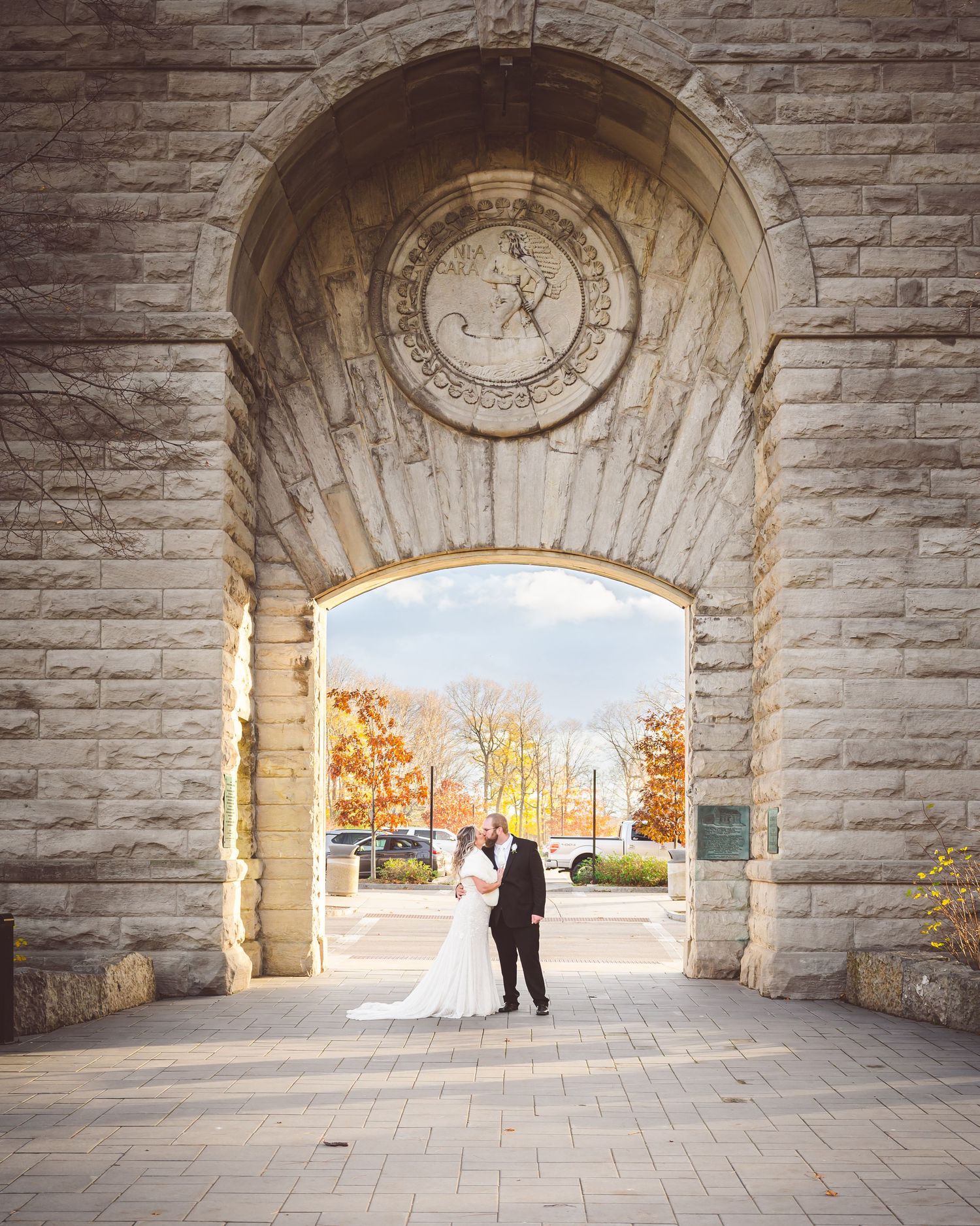 Newlyweds stand under a stone archway during autumn with trees in the background.