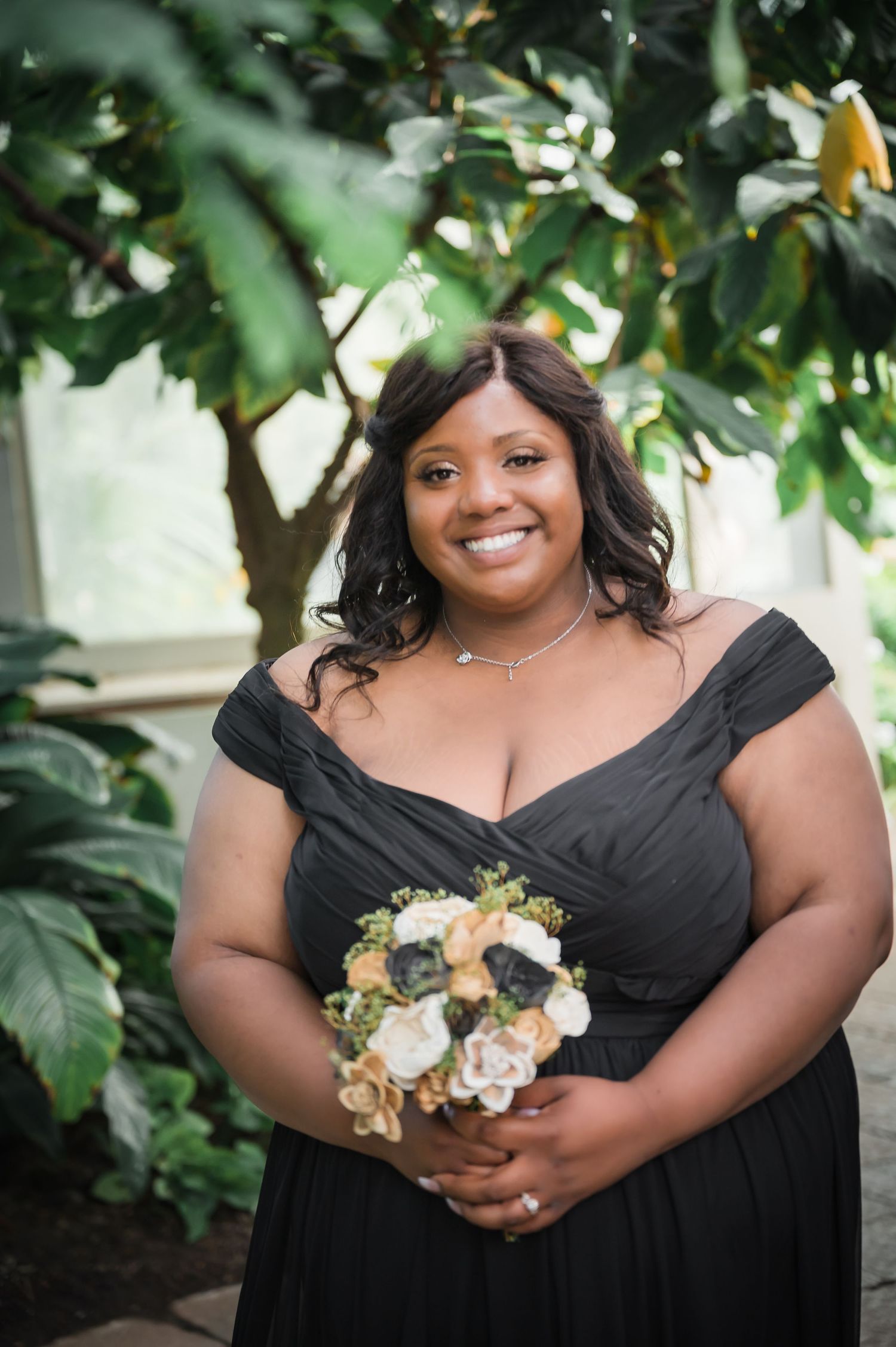 Bridesmaid in black dress holding peach and white flower bouquet poses in garden setting.