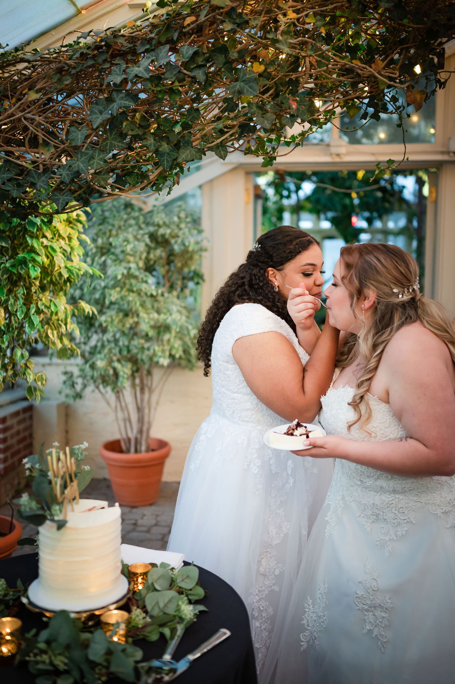 Two brides in white wedding dresses share cake at an outdoor reception with greenery and a white cake.