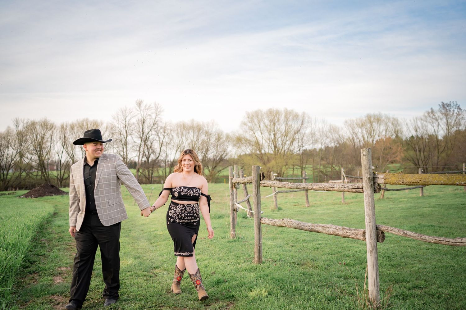 A couple walks hand in hand through a rustic farm pasture with wooden fencing.