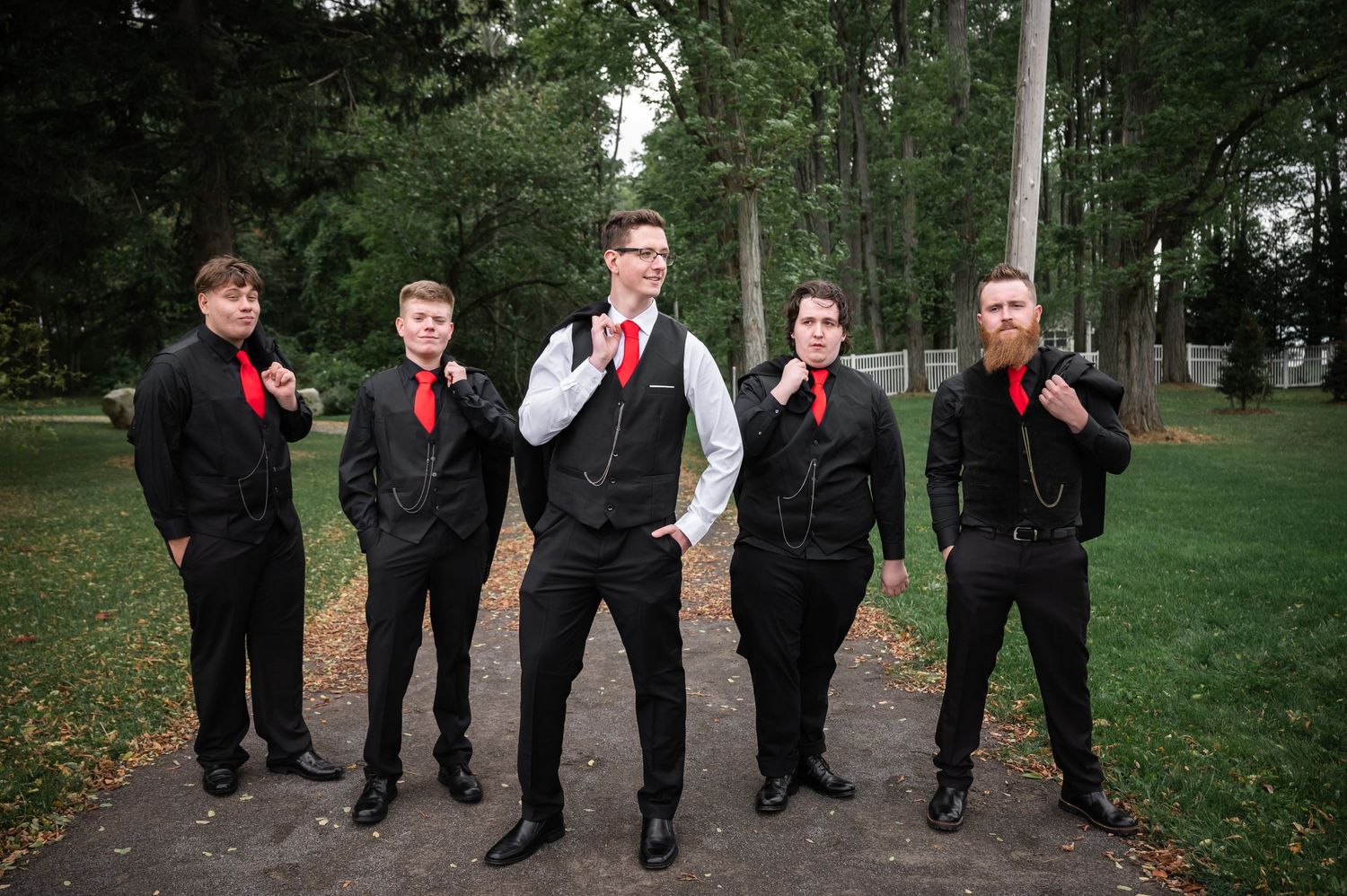 Groomsmen in black suits with red accents pose casually on a tree-lined pathway.