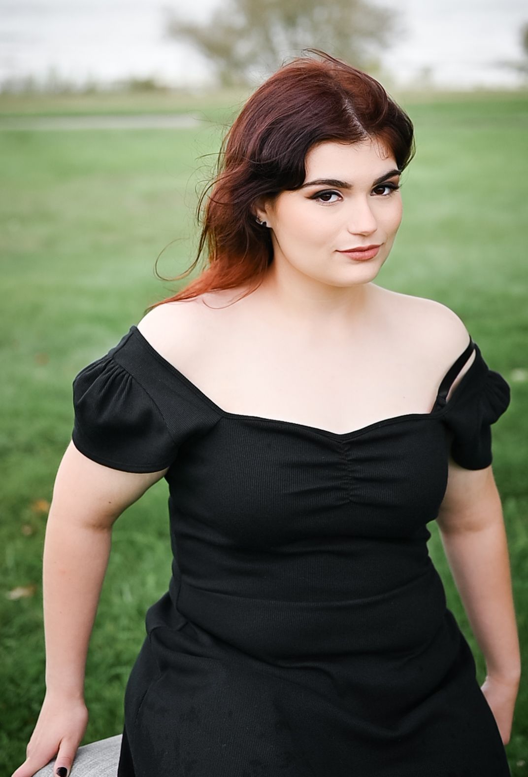 A portrait outdoors wearing a black off-shoulder dress against a green grassy background.