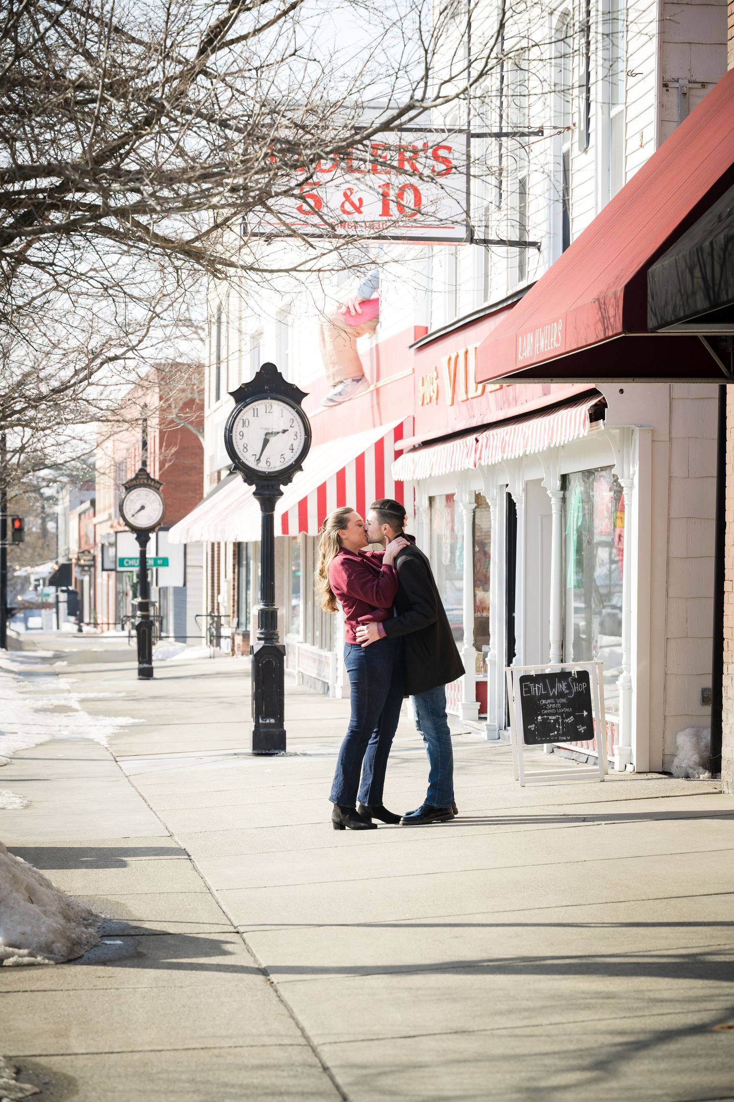 Couple sharing a romantic kiss on a snowy small-town sidewalk beside a vintage street clock and red-awning shops.