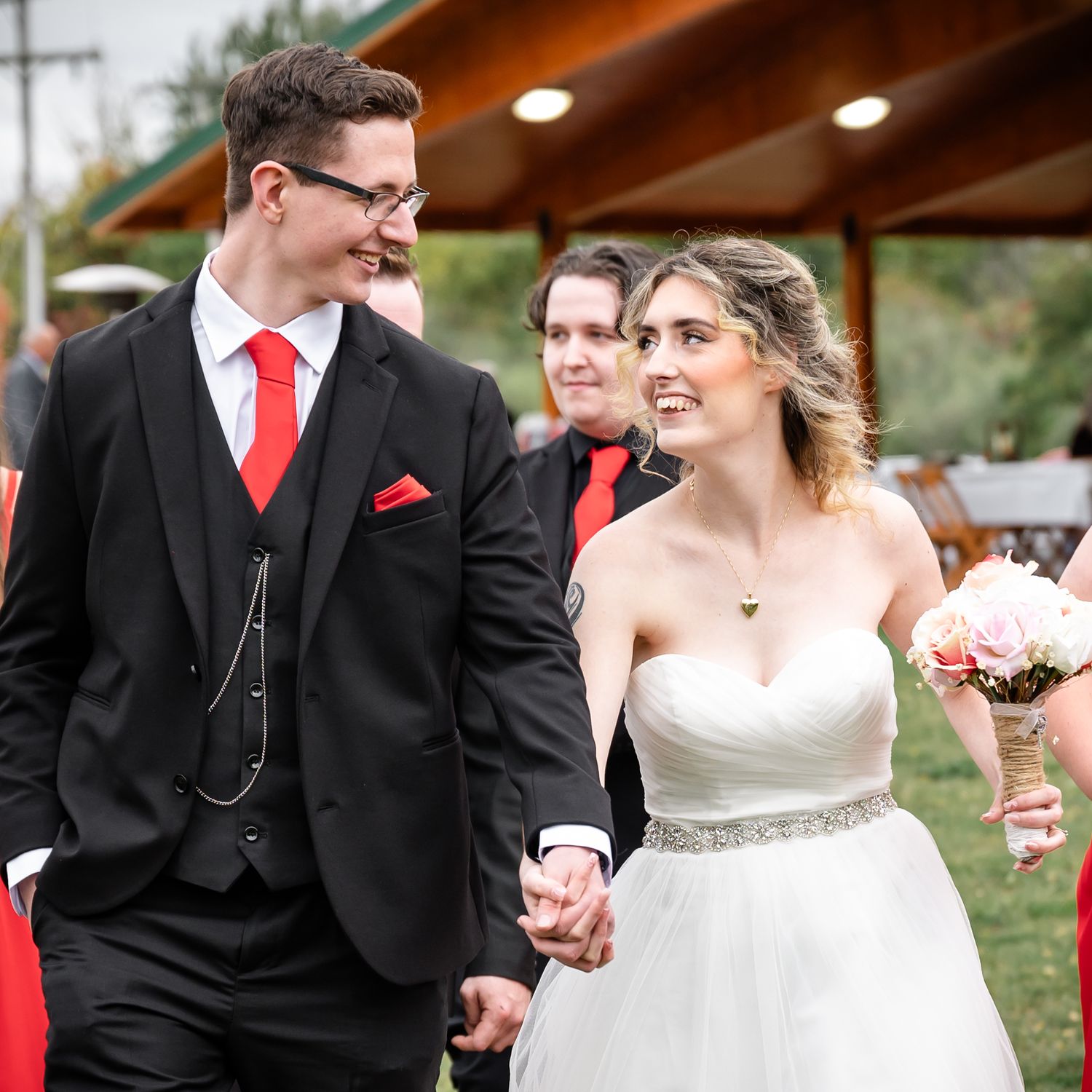 Newlyweds hold hands while walking outdoors near wooden pavilion.
