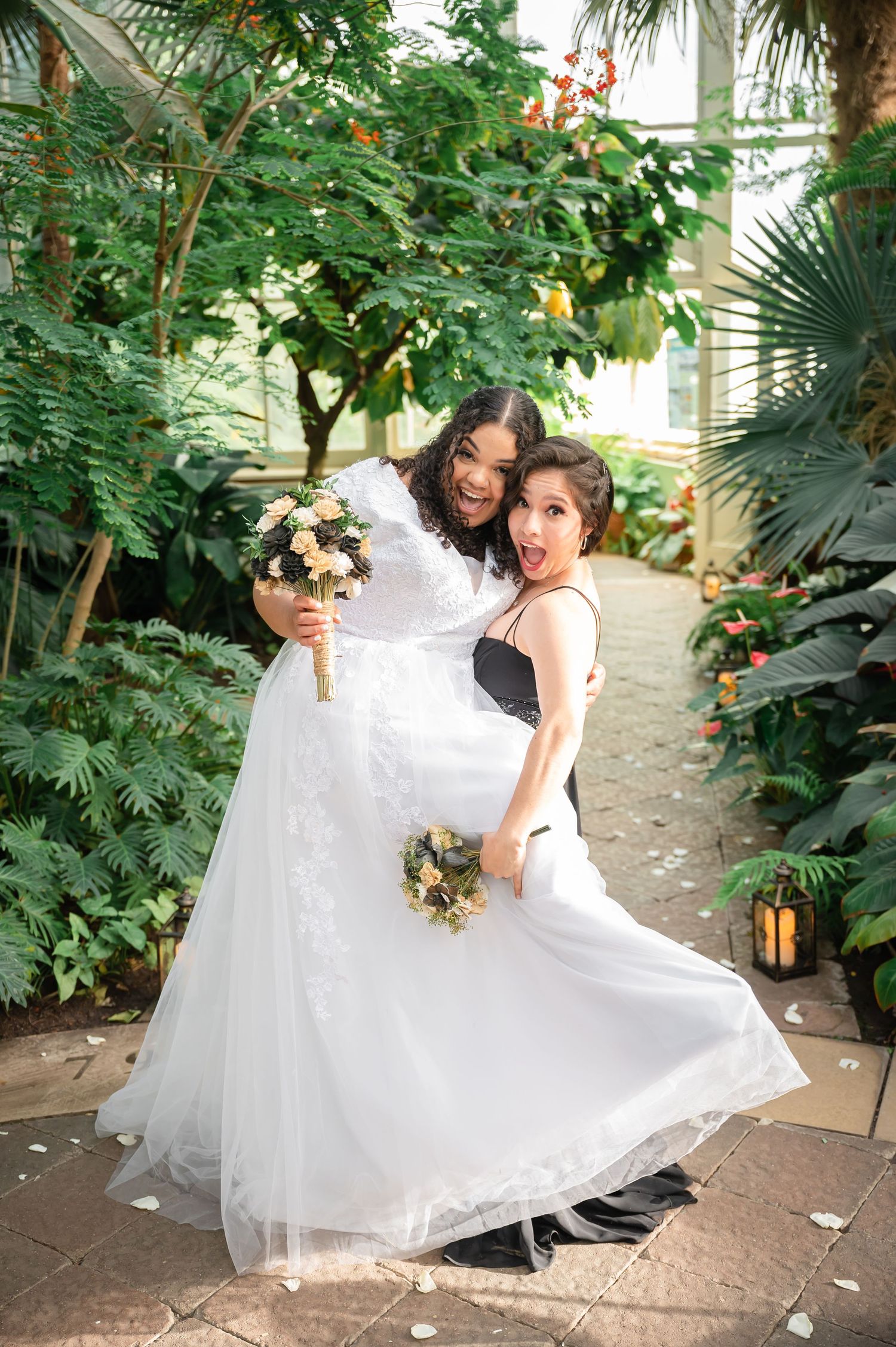 Two people in white dresses pose playfully in a tropical garden setting with bouquets.