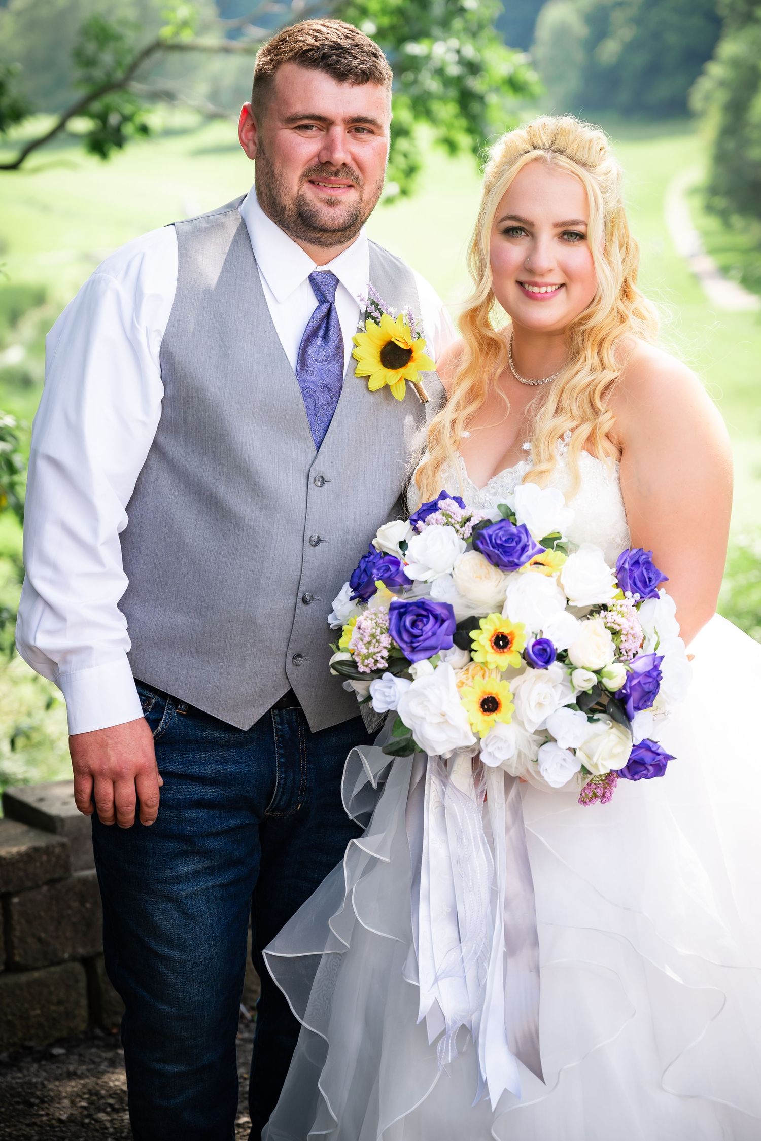 Wedding couple poses outdoors with sunflower and purple floral bouquet against natural landscape.