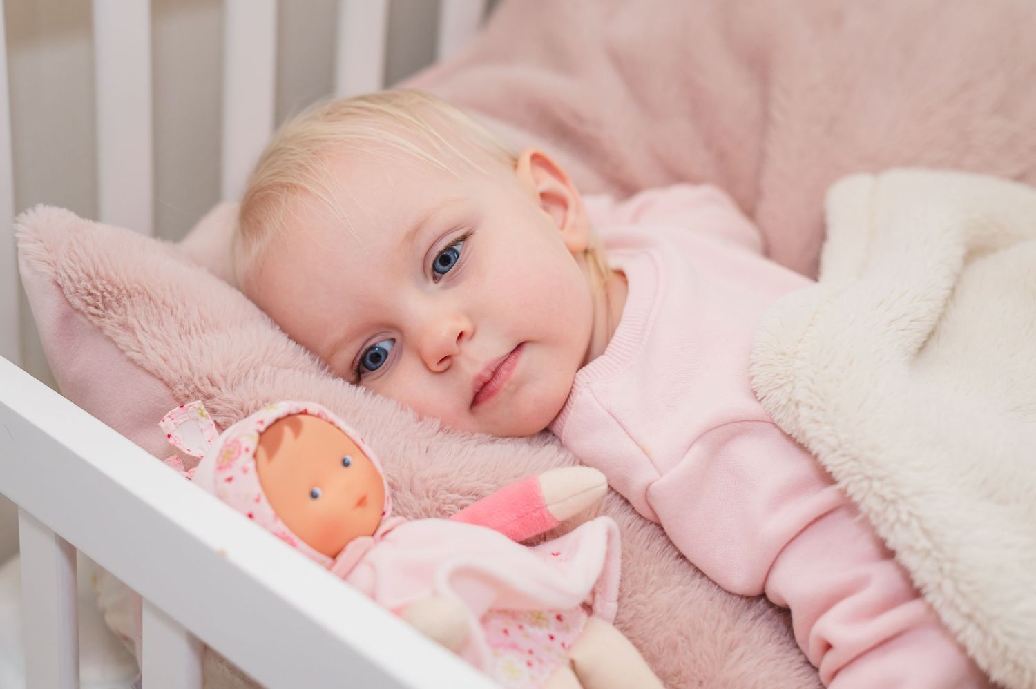 Child resting in white crib with pink clothing and plush doll.