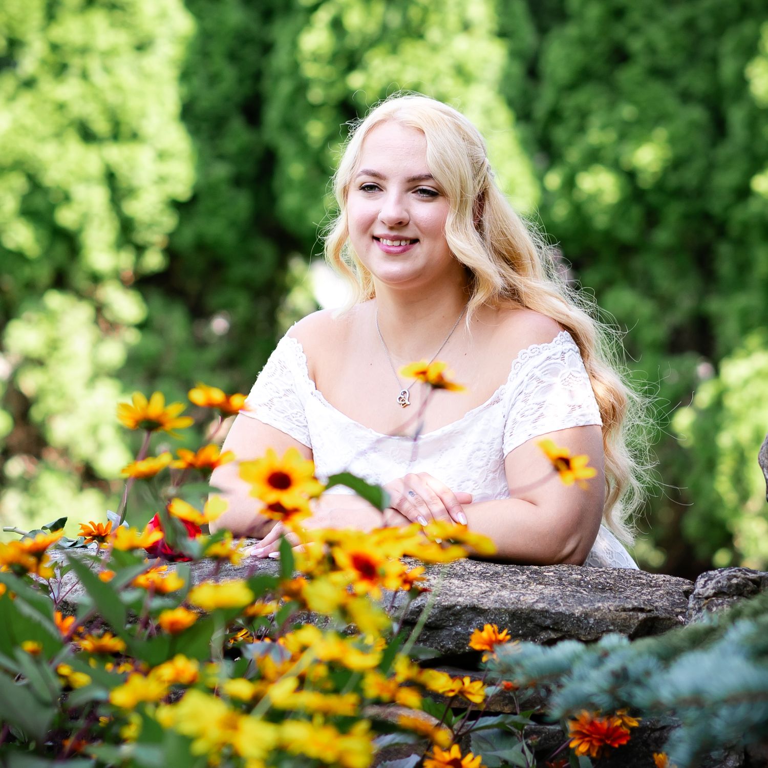 Woman in white dress poses by stone wall surrounded by bright yellow and orange flowers.