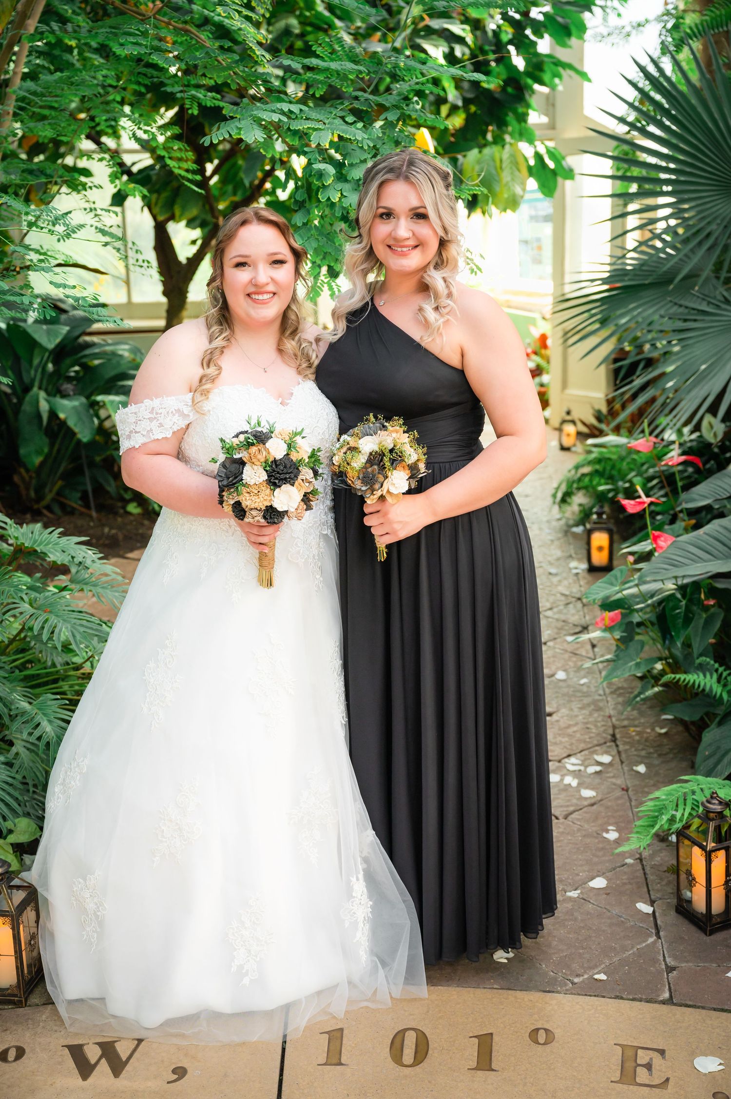 A bride in white and bridesmaid in black hold sunflower bouquets on a garden path.