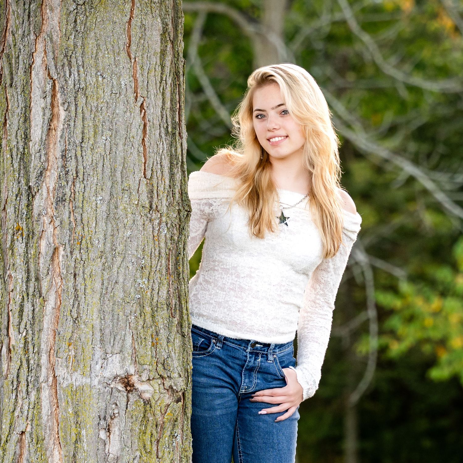 A person in a white off-shoulder sweater and jeans poses casually against a large tree trunk outdoors.