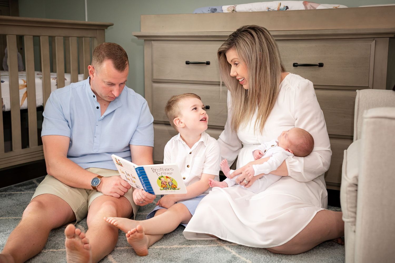 Family sits together on floor of nursery reading a book while holding newborn baby.