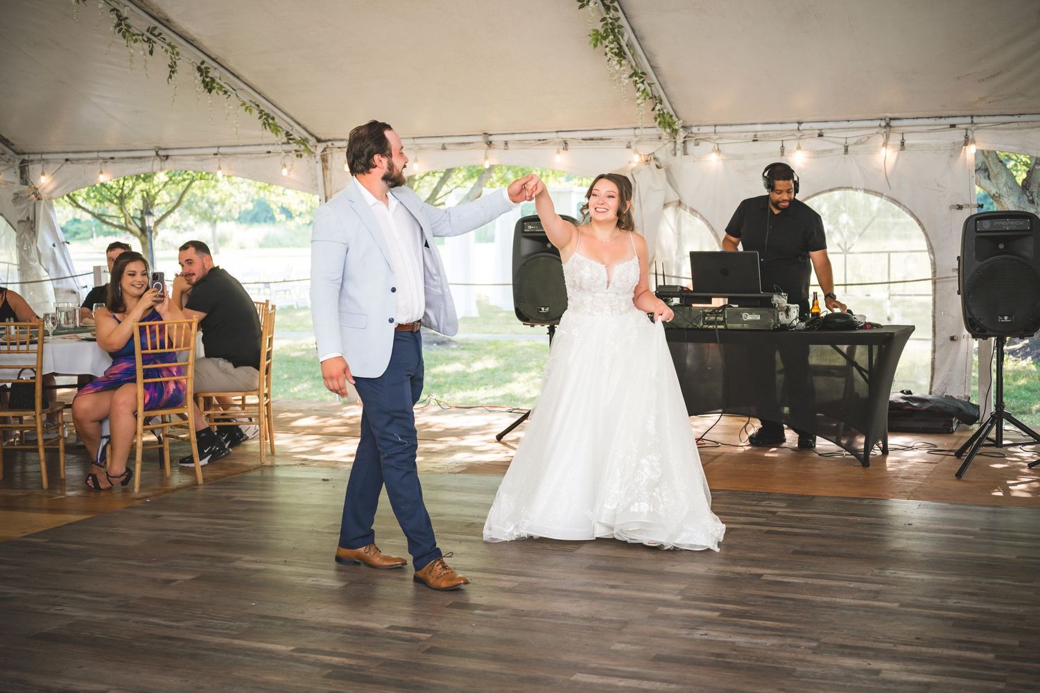 A couple shares their first dance at an outdoor wedding reception under a white tent with guests and a DJ in the background.