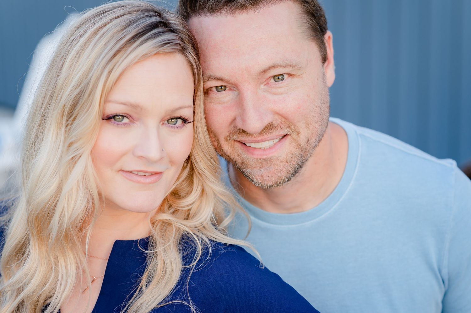 A happy couple poses together outdoors with light blue wall behind them, wearing casual navy and light blue tops.