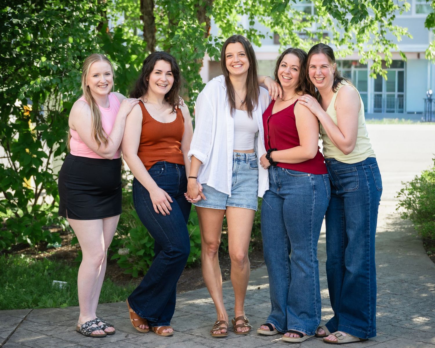 Five young women smiling and posing together outdoors on a sunny day near green trees and a building.